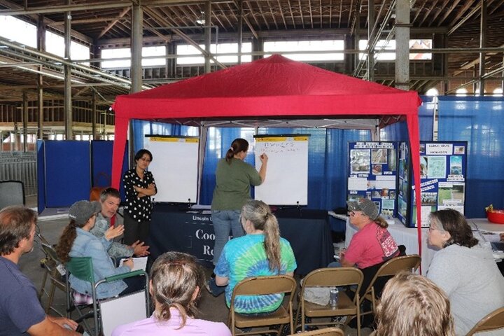 Group sitting in chairs under a red tent, watching a presenter write on a large easel.
